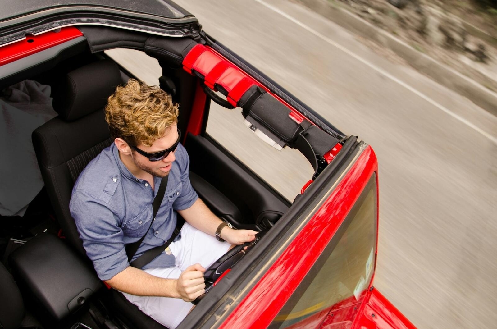 Man driving a red CJ Jeep with the top off, hand on wheel, late afternoon sun
